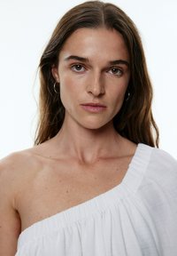 Haut blanc à épaules dénudées avec texture plissée, une seule manche, en tissu léger et à coupe décontractée; le mannequin a de longs cheveux bruns.