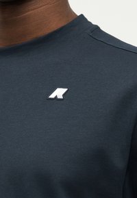 Κοντινό πλάνο σκούρου μπλε πουκαμίσου που φοριέται από ένα άτομο, με ένα μικρό λευκό και μαύρο γεωμετρικό λογότυπο στο στήθος κοντά στον γιακά.