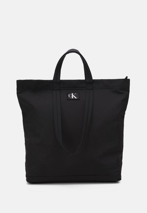 Zwarte draagtas met dubbele handvatten, Calvin Klein-logo op de voorkant en bovenste ritssluiting op een effen achtergrond.