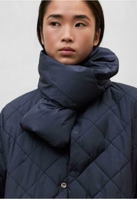 Chaqueta de plumas acolchada en color azul marino con un gran cuello enrollado. Presenta botones a presión y una textura suave y brillante. Diseño elegante y moderno.