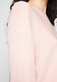 Femme portant un pull en maille rose pâle avec un col rond et un tissu texturé, visible des épaules au cou.