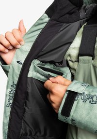 Zielona zewnętrzna kurtka o teksturowanym wykończeniu, z czarną podszewką, regulowanymi mankietami oraz wewnętrzną kieszenią zapinaną na rzep.