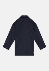 Navy blue jacket with long sleeves, collared neck, buttoned cuffs, and a smooth texture. Features a circular logo on the lower hem.