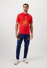 Crvena majica s zlatnim grbom španjolske reprezentacije, uparena s tamnoplavim hlačama s zlatnim detaljima i logotipom. Ležerna sportska odjeća.