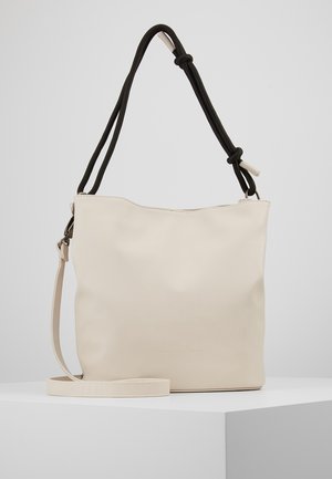 Sac cabas crème avec bandoulière beige réglable et poignée d’épaule noire nouée, présenté sur un socle blanc sur fond neutre.