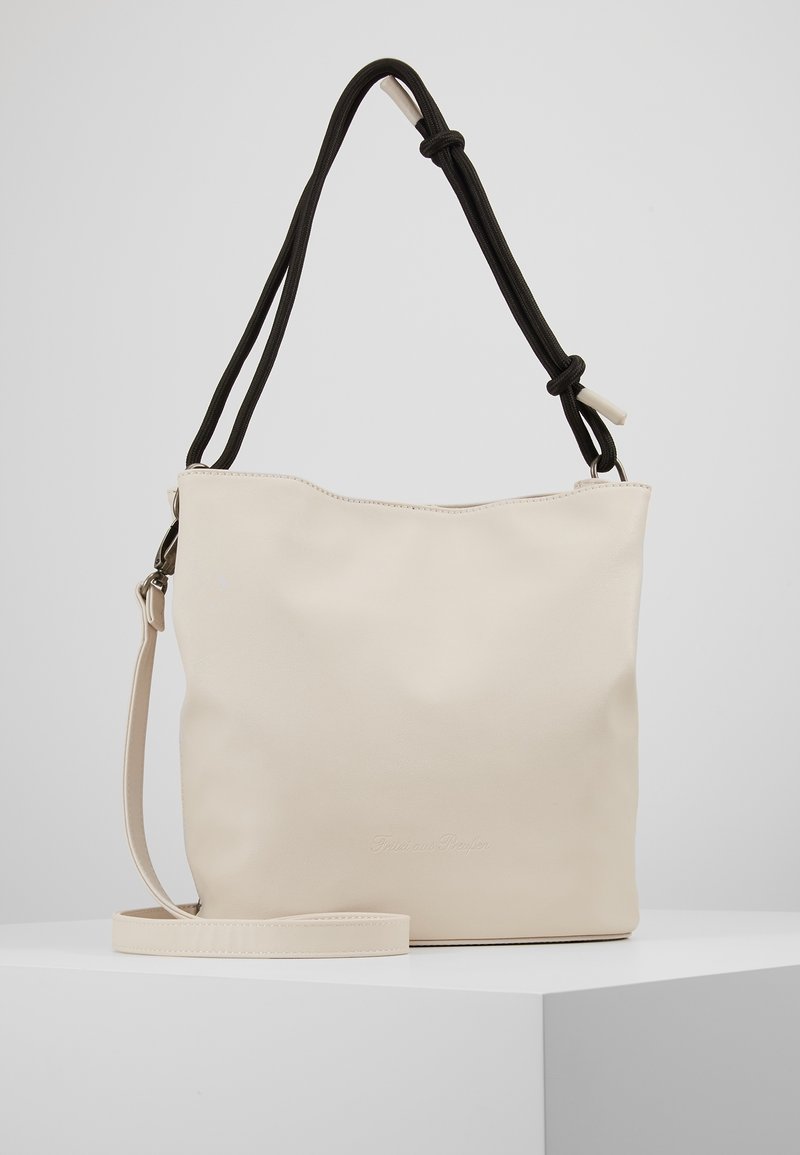 Sac cabas crème avec bandoulière beige réglable et poignée d’épaule noire nouée, présenté sur un socle blanc sur fond neutre.