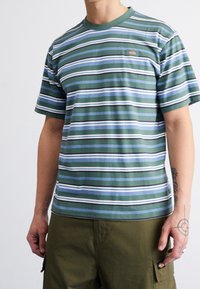 Muž v zeleném, modrém, černém a bílém pruhovaném tričku a olivových cargo kalhotách, s tetováními viditelnými na levém předloktí a ruce.