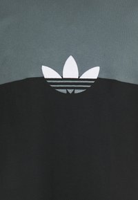 Camiseta negra y gris con un gran logo blanco de trébol de Adidas; tejido texturizado, cuello redondo, diseño en bloques de color.