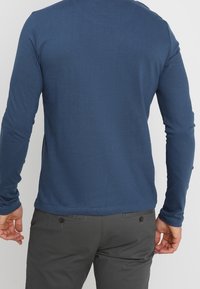 Tmavě modrý top s dlouhými rukávy z bavlny, s kulatým výstřihem a přiléhavým střihem. Nošený s šedými kalhotami, s minimem švů a detailů.