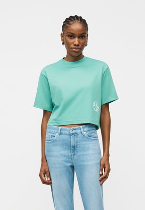 Mladá žena v mentolovo zelenej krátkej tričku s malým bielym logom a svetlomodrých nohaviciach s vysokým pásom, stojaca pred jednoduchým pozadím.