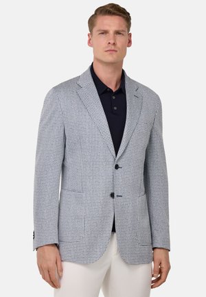 Blazer a quadri blu chiaro e bianco realizzato in tessuto testurizzato, con rever a punta, due bottoni e tasche frontali. Indossato sopra una camicia scura.