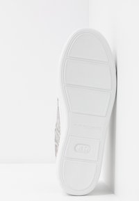 Podešev bílé tenisky s texturovaným vzorem protiskluzu, boční pohled ukazuje světle hnědé a šedé tóny, s logem značky na podrážce.