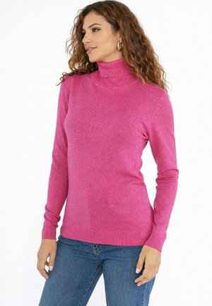 Femme aux cheveux bouclés portant des boucles d'oreilles créoles dorées, un pull à col roulé rose vif et un jean bleu, debout devant un fond blanc.