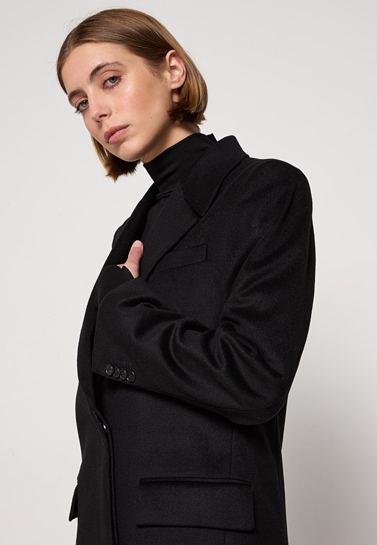Femme aux cheveux courts portant un col roulé noir et un blazer noir surdimensionné, tenant le revers du blazer avec une expression neutre.