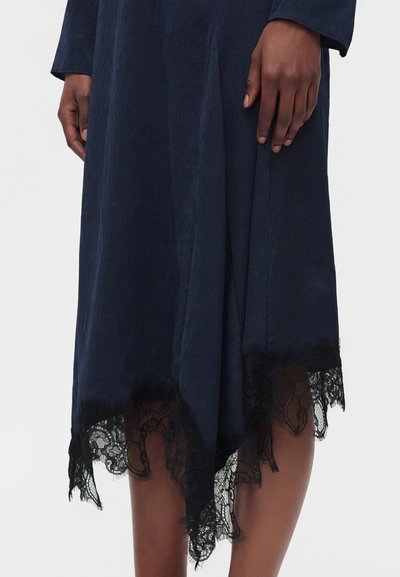 Femme portant une robe bleu marine foncé en tissu texturé avec un ourlet asymétrique orné d'une bordure en dentelle noire, les mains reposant le long du corps.