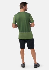 Grön kortärmad träningströja med texturerad tyg, svarta detaljer, i kombination med svarta shorts och mörka skor. Vy från baksidan.