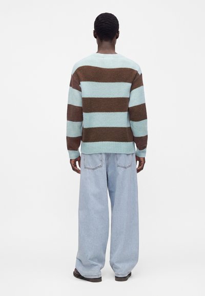 Homme vu de dos portant un pull à larges rayures marron et bleu clair, un jean ample bleu clair et des chaussures foncées sur fond blanc.