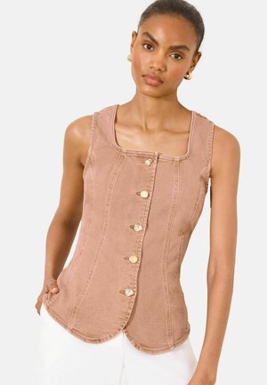 Donna che indossa un top marrone senza maniche con bottoni e orlo smerlato, e pantaloni bianchi, in piedi contro uno sfondo chiaro e uniforme.