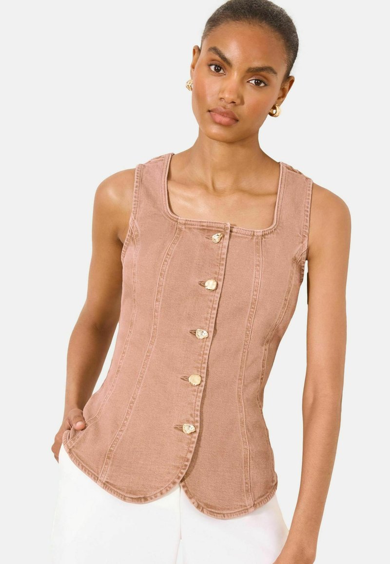 Woman wearing sleeveless, buttoned brown top with scalloped hem and white pants, standing against plain light background.