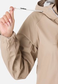 Veste imperméable beige avec capuche à cordon de serrage. Comprend des poignets élastiques, une texture lisse et des détails de surpiqûres visibles.