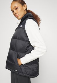 Μαύρο γιλέκο τύπου πούφας με πλαϊνές τσέπες, ψηλό γιακά και γυαλιστερή υφή. Διαθέτει έντονο λογότυπο και σήμανση 700 fill power.