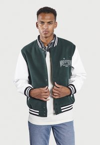 Chaqueta varsity de mezcla de lana verde con mangas blancas, puños a rayas en blanco y negro, detalle en letras en el frente y cierre de botones a presión.