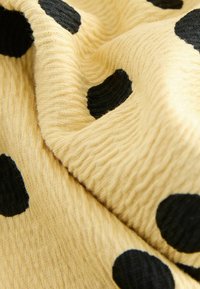 Tela amarilla con una superficie texturizada, con grandes lunares negros de varios tamaños, drapeada para mostrar su suavidad y flexibilidad.