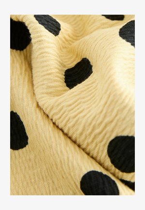 Geltonos spalvos audinys su tekstūruota paviršiaus struktūra, puoštas didelėmis juodomis spalvos taškeliais įvairių dydžių, permestu taip, kad matytųsi jo minkštumas ir lankstumas.