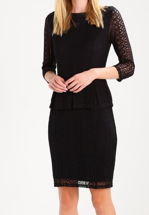 Frau trägt ein schwarzes, figurbetontes Spitzenkleid mit dreiviertel durchsichtigen Ärmeln und Peplum-Taillendetail, steht mit verschränkten Händen.