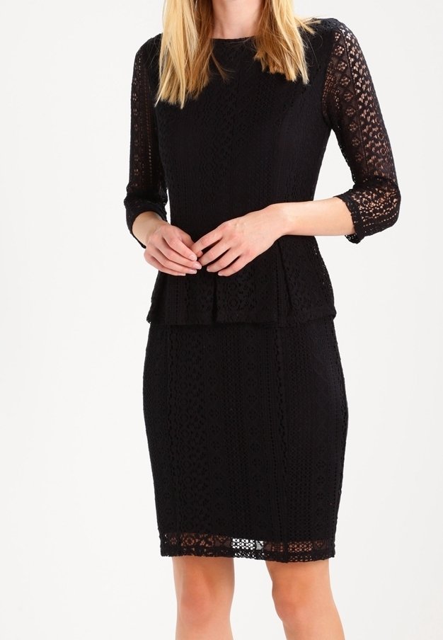 Femme portant une robe noire ajustée en dentelle avec des manches trois-quarts transparentes et un détail peplum à la taille, debout les mains jointes.