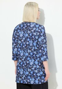 Bluse mit blauem Blumenmuster und Dreiviertelärmeln, auf einem dunklen Hintergrund aus weichem Stoff. Sie zeigt eine fließende Silhouette und einen abgerundeten Saum.