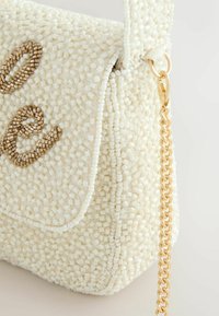 Sac à main blanc perlé avec anse chaîne dorée et lettrage décoratif en perles marron sur le rabat avant, vue en gros plan.