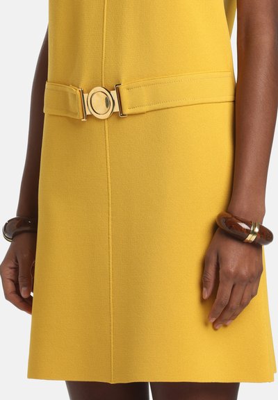 Rochie galbenă cu o curea aurie cu cataramă circulară la talie, purtată împreună cu două brățări groase maro și aurii pe ambele încheieturi.