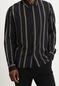 Zwarte blouse met lange mouwen, voorzien van verticale witte en gouden strepen, knoopsluiting en een klassieke kraag. Gladde stofstructuur.