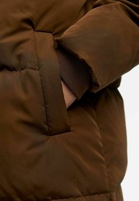 Giacca piumino marrone con sezioni trapuntate e una tasca laterale. Presenta un polsino interno in tessuto marrone strutturato. Materiale morbido e isolante.
