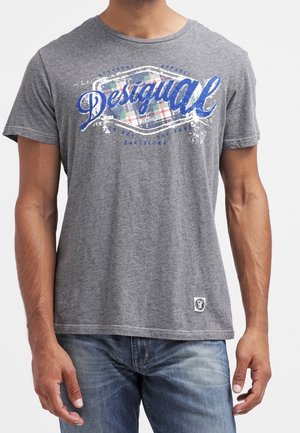 Homme portant un t-shirt gris Desigual avec un texte graphique bleu et blanc, et un jean bleu clair, debout devant un fond uni.