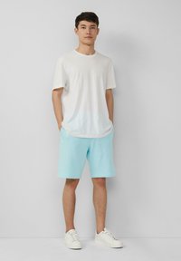 Männliches Outfit bestehend aus einem weißen Baumwoll-T-Shirt und hellblauen Shorts. Die Shorts haben einen elastischen Bund und Taschen. Abgerundet mit weißen Sneakers.