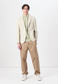 Blazer texturé beige sur une chemise vert clair, associé à un pantalon kaki et des sneakers blanches. Le blazer présente une fermeture à un bouton et des poches.