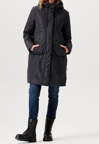 Parka negra con capucha y cierre de cremallera, que cuenta con dos bolsillos laterales, un diseño acolchado y un corte relajado. Combinada con jeans oscuros y botas negras.