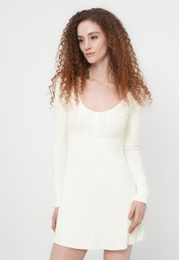 Λευκό πλεκτό φόρεμα με μακριά μανίκια, στρογγυλή λαιμόκοψη και υφή πλέξης με καλώδια. Διαθέτει εφαρμοστό μπούστο και φαρδιά πλάγια.