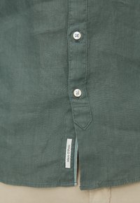 Zielona lniana koszula z prostym dołem, małym bocznym rozcięciem i dwoma białymi guzikami. Na dole po lewej stronie znajduje się metka z napisem "Marc O'Polo".