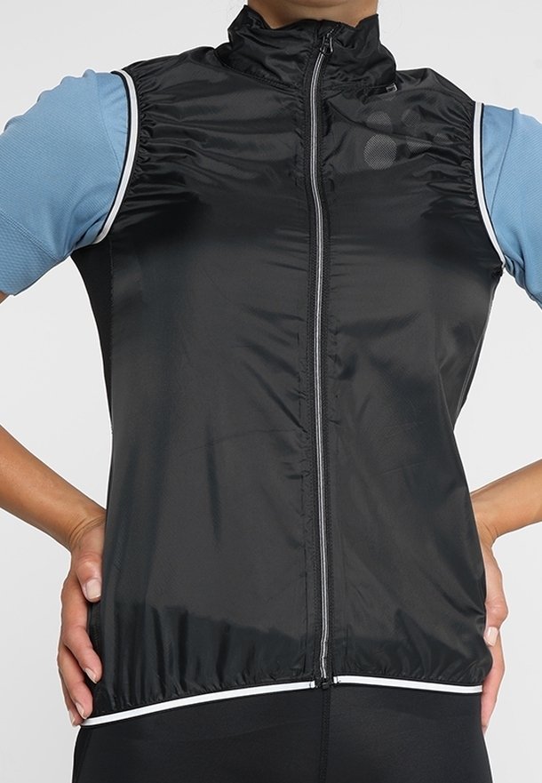 Personne portant une veste noire sans manches zippée sur une chemise bleu clair à manches courtes, debout les mains sur les hanches.