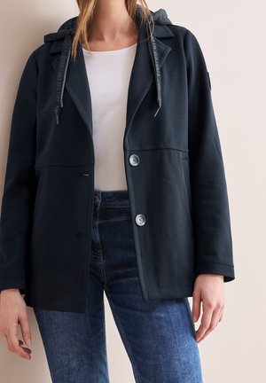 Personne portant une veste noire boutonnée avec une capuche par-dessus une chemise blanche et un jean bleu, debout contre un mur uni.