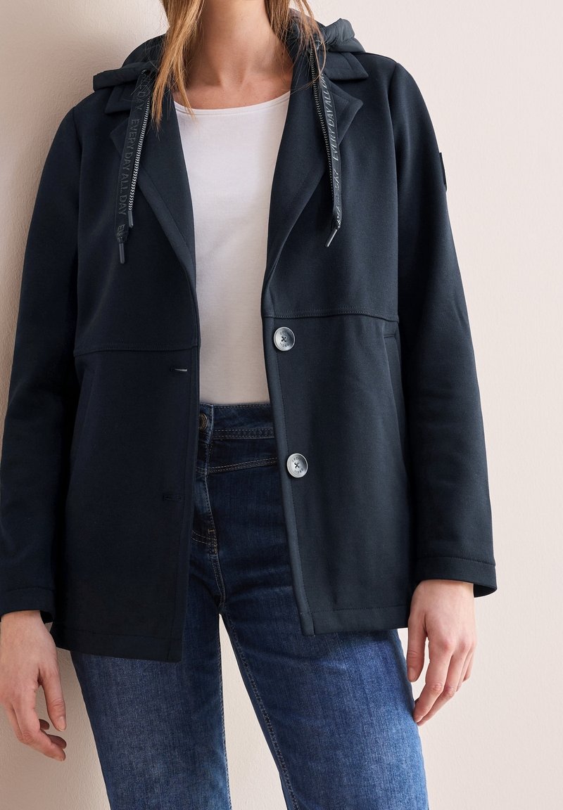 Personne portant une veste noire boutonnée avec une capuche par-dessus une chemise blanche et un jean bleu, debout contre un mur uni.