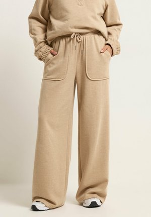 Person iført beige sweatpants med vide ben, forlommer og snøre i taljen, kombineret med hvide sneakers, stående mod en ensfarvet baggrund.