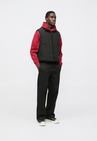 Zwarte gewatteerde bodywarmer over een rood met capuchon sweatshirt, gecombineerd met zwarte broek en zwarte sneakers met witte zolen en accenten.