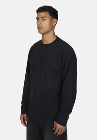 Jonge man in een zwart longsleeve t-shirt met ronde hals en subtiele geborduurde logo, staande tegen een simpele lichte achtergrond.