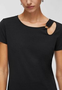 Schwarzes Kurzarmoberteil mit geschwungenem Ausschnitt und betonter Aussparung an der Schulter, die durch ein Metallringdetail gesichert ist. Weiche Stofftextur.