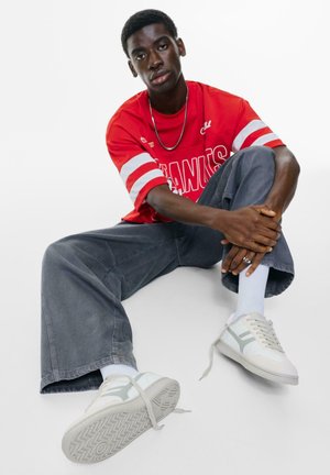 Rotes Netz-Sporttrikot mit weißen Streifen, graue weite Jeans und weiße Sneaker mit grauen Akzenten, auf einem weißen Hintergrund sitzend.