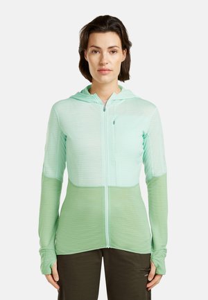 Vrouw draagt een mintgroen gewatteerd jack met capuchon en duimgaten, staand tegen een effen witte achtergrond.